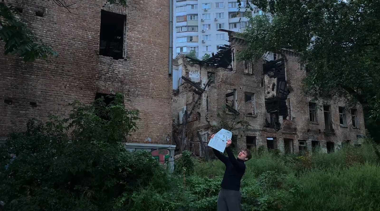 Holding up paper in front of a ruined building, music video still